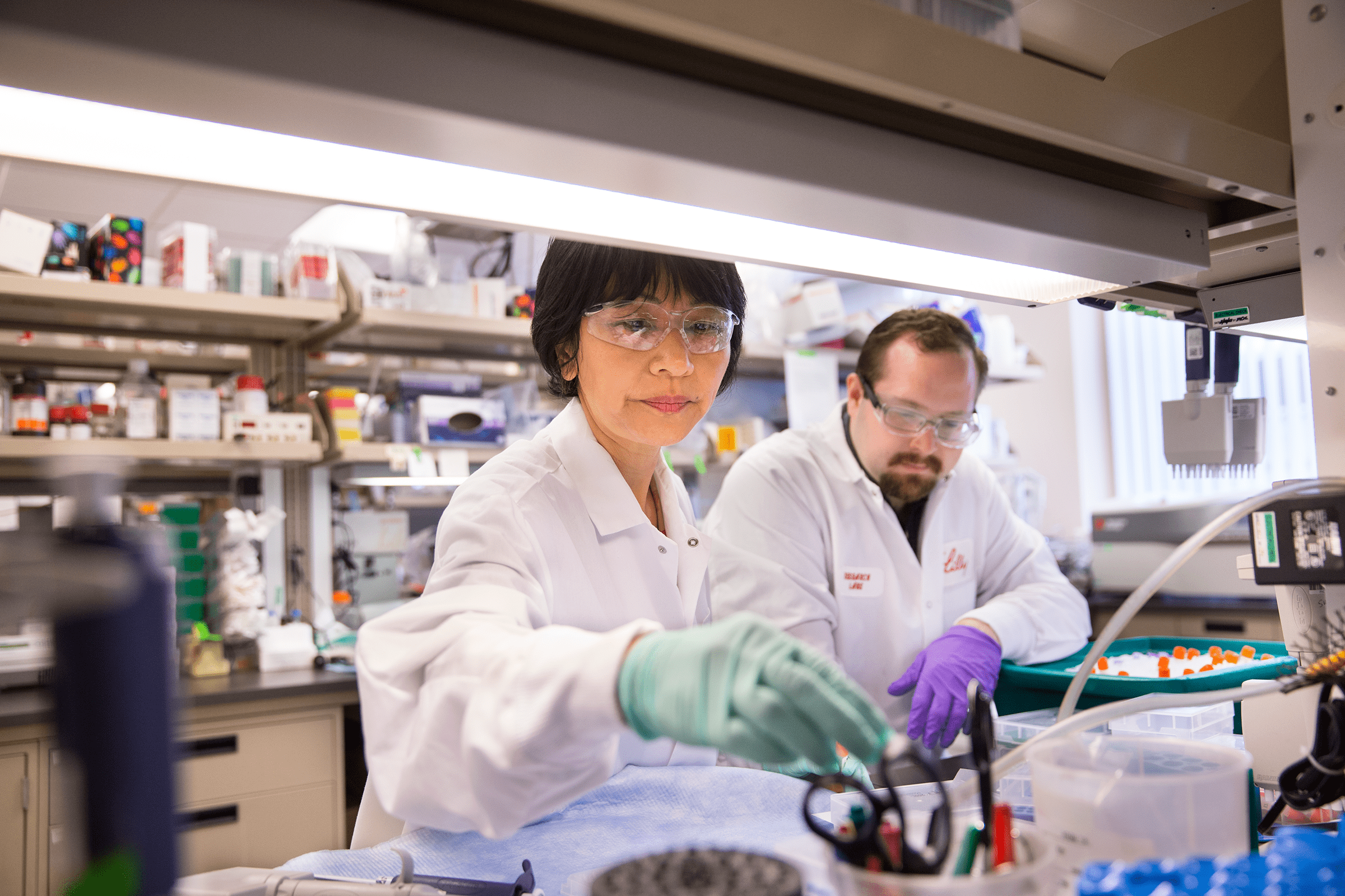 male and female scientists wearing white Lilly lab coats and gloves working in lab